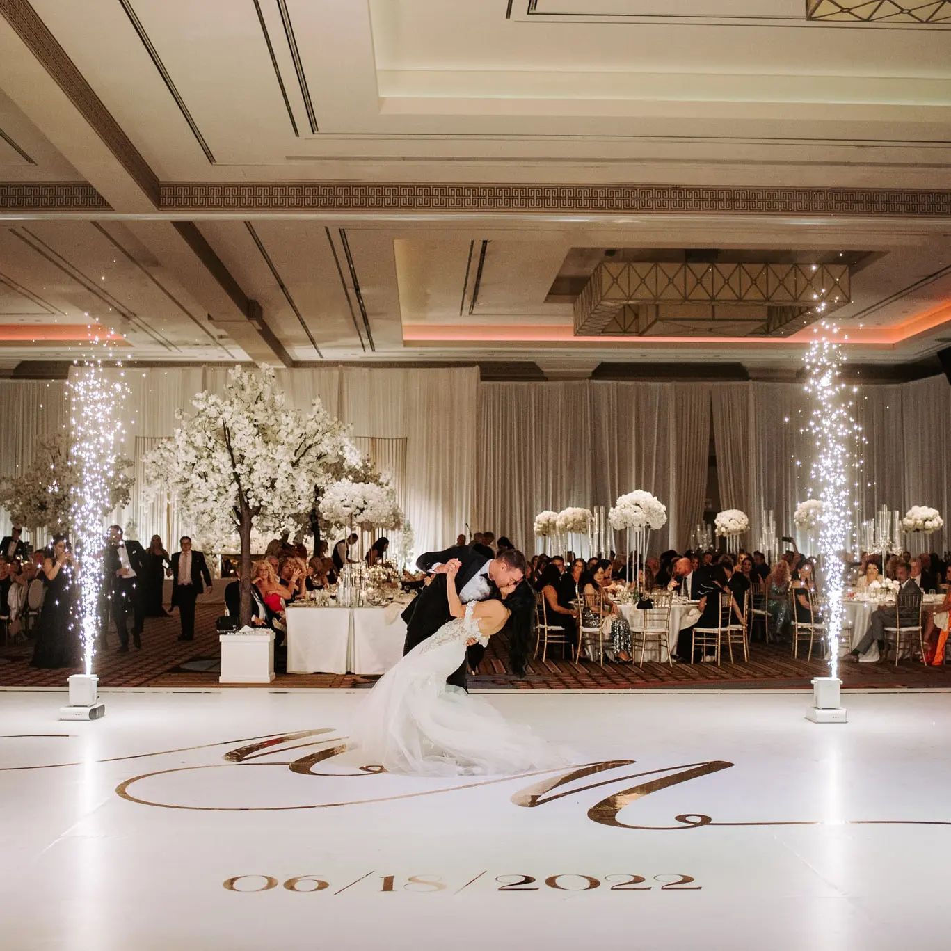 A bride and groom mid-first-dance on a custom monogrammed white vinyl floor wrap printed with their wedding date, indoor sparkler fountains framing the moment.